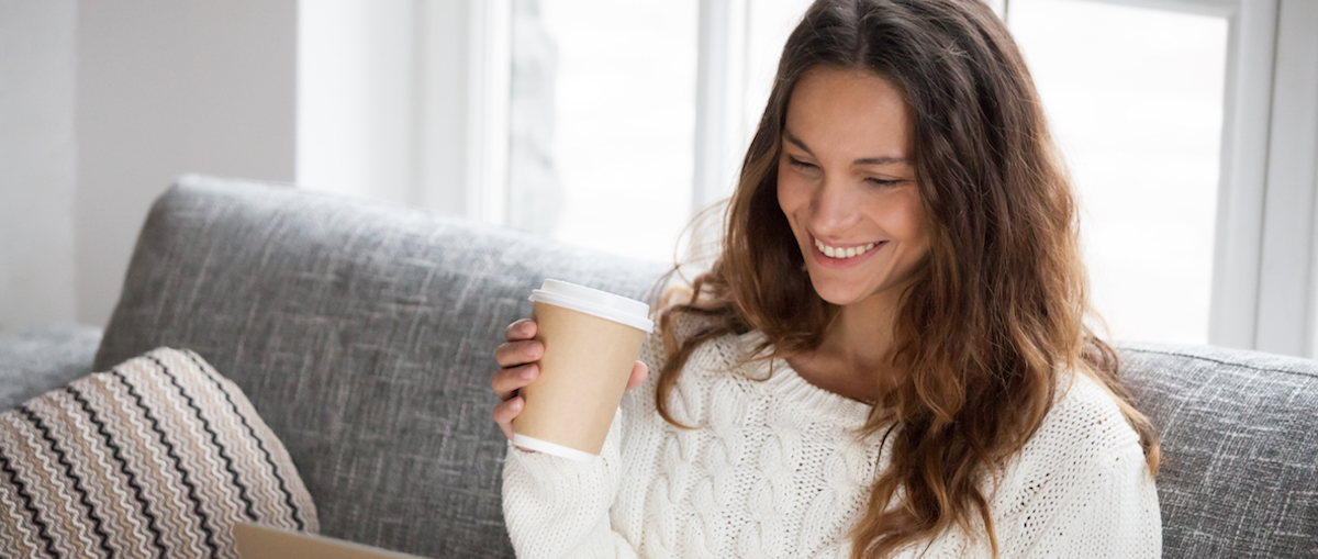 Smiling young woman enjoying morning coffee using laptop sitting at home on sofa, attractive happy girl  typing on computer having fun online or chatting with friends while relaxing on couch