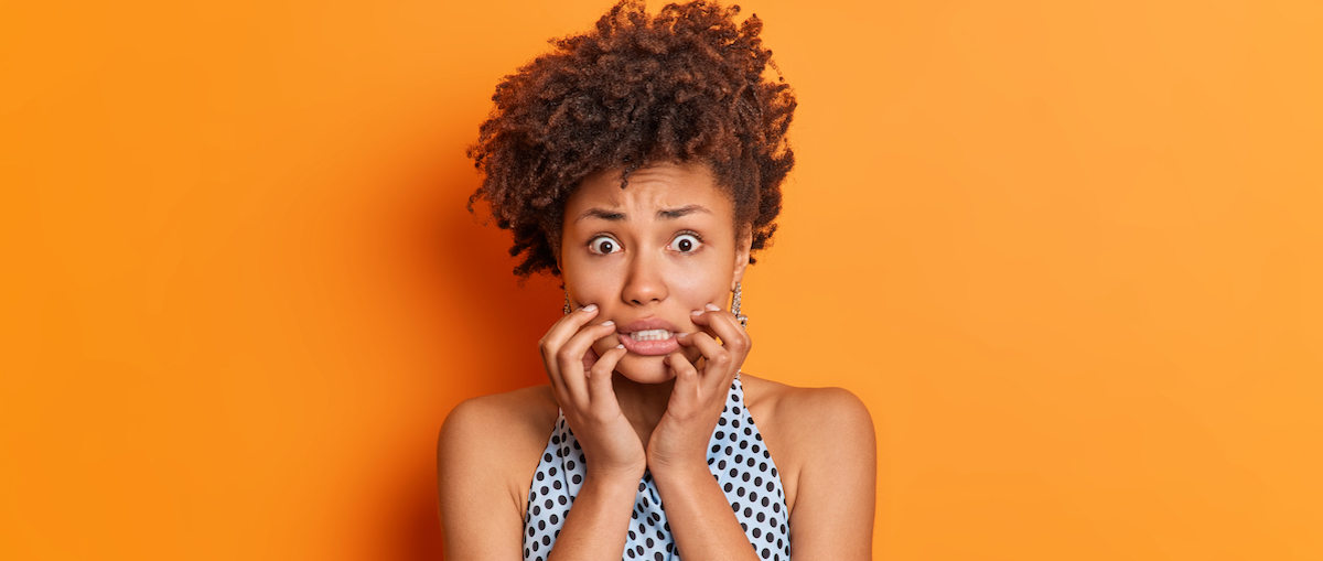 Nervous worried Afro American woman clenches teeth looks embarrassed at camera wears fashiobnable outfit afraids of bad consquences poses in studio against orange background. Scared female model